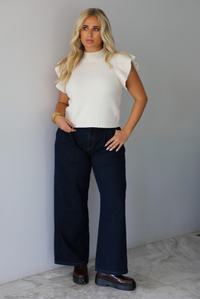 A woman wearing a cream ribbed knit sweater top with ruffled sleeves, dark wide-leg denim jeans, and brown loafers, standing indoors against a white wall with white marble flooring. 