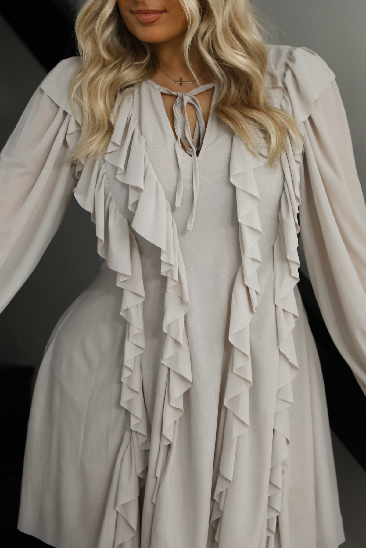 Woman model wearing a light taupe romper that looks like a dress. The photo is taken indoors against a white wall with a black railing and black steps. The photo is close up and shows the ruffle details in the front. 