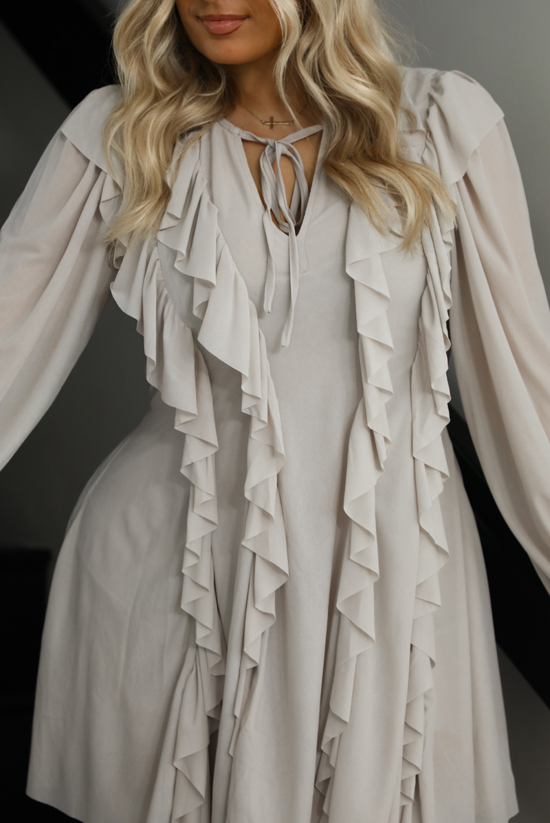 Woman model wearing a light taupe romper that looks like a dress. The photo is taken indoors against a white wall with a black railing and black steps. The photo is close up and shows the ruffle details in the front. 