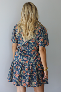 A woman shown from the back modeling a short-sleeve floral mini dress with a tie waist, standing indoors against a white wall.