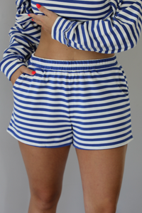 Woman wearing a navy and white striped two piece set featuring a long sleeved top and shorts indoors against a white wall. The photo shows a closeup of the shorts, which have an elastic waistline and functional pockets on either side. 
