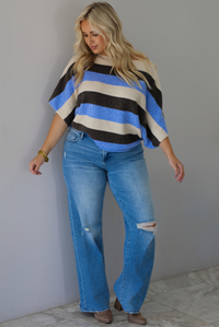 A woman wearing a cream, brown,, and blue striped knit top that has a boat-neck design, and quarter length flowy sleeves, paired with a pair of medium wash denim jeans, standing indoors against a white wall with white marble flooring. 