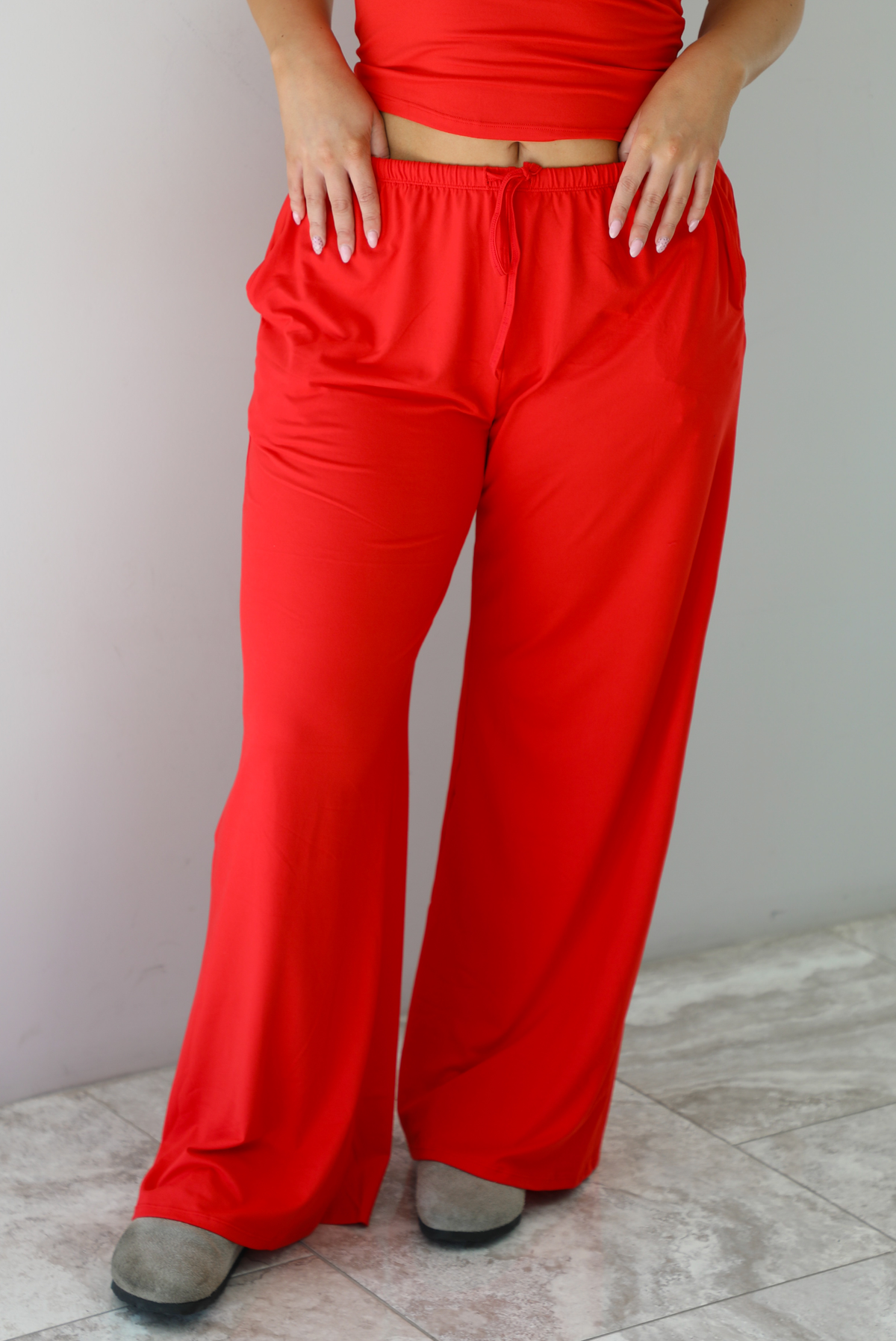 Woman wearing a red two piece set featuring a  tank top and slouchy pants. The photo shows a closeup of the pants. The photo is taken indoors against a white wall with white marble flooring. 
