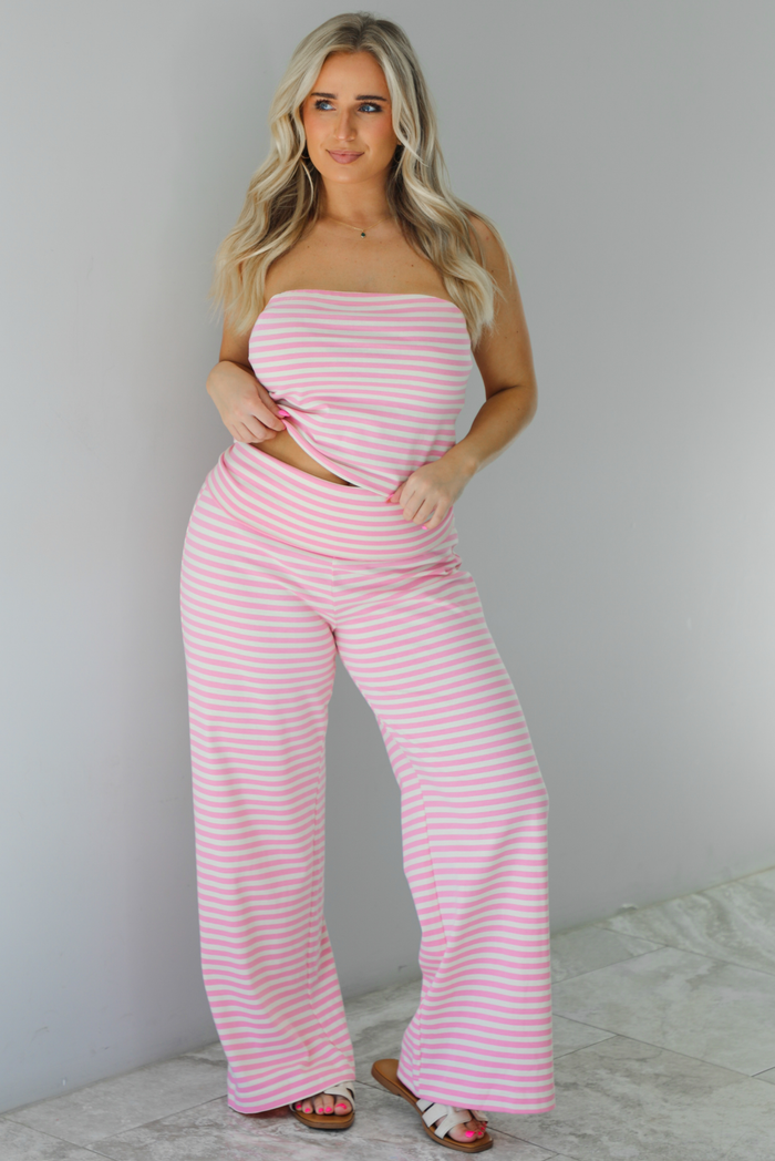 Woman wearing a pink and cream striped two piece set featuring a tube top strapless top and rolled-over waistline, flared hem pants. The photo is taken indoors against a white wall with white marble flooring. 