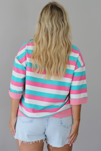 Woman wearing turquoise, pink, and white striped top with light wash denim shorts indoors against a white wall. The photo shows the back of the top. 