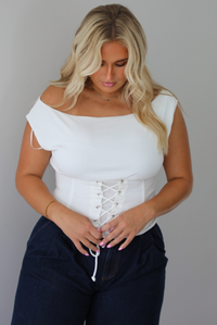 A woman modeling a white off-the-shoulder lace-up corset top paired with dark denim wide-leg jeans and pinkish-brown flat shoes, standing indoors against a white wall.