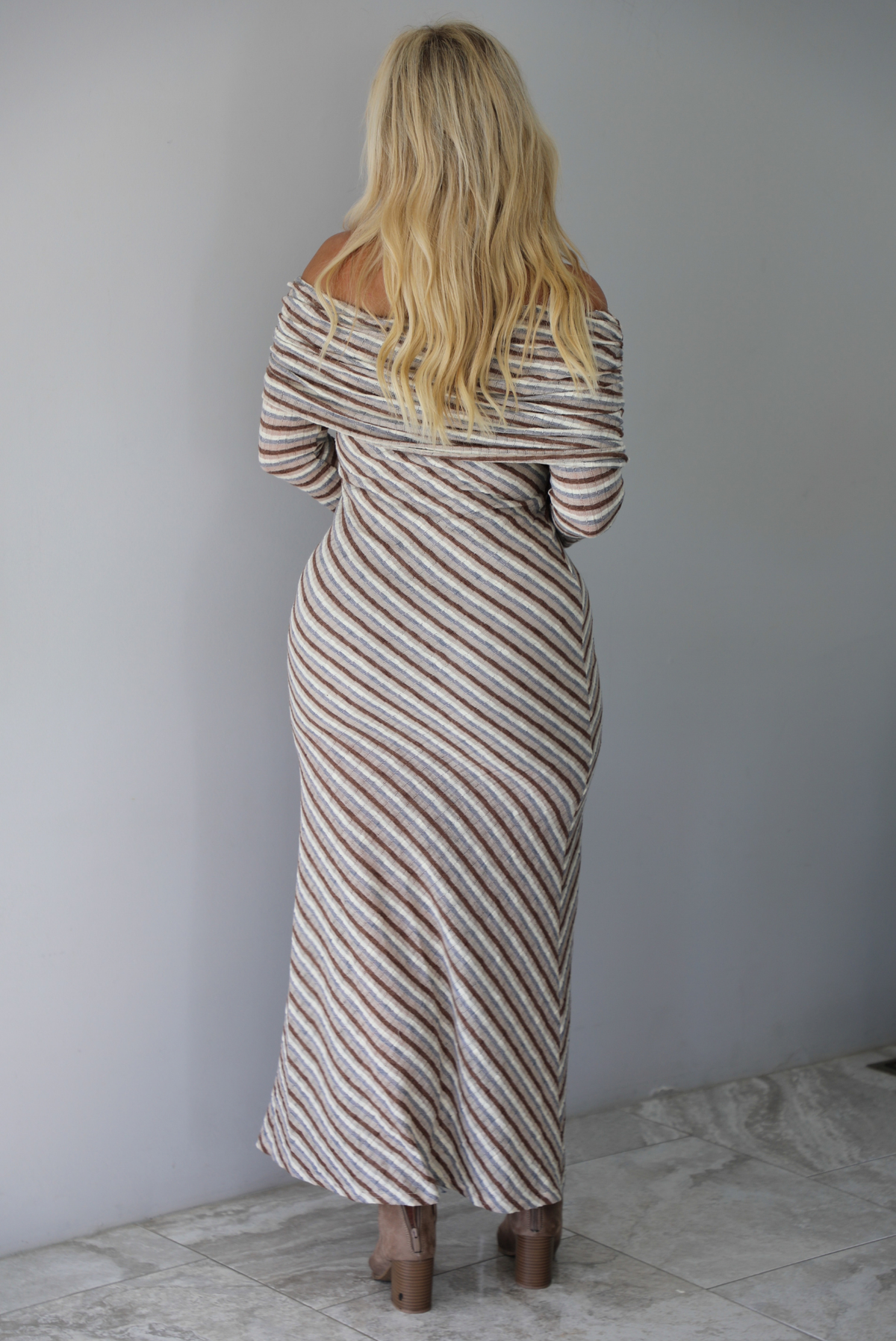 A woman shown from behind wearing a grey, brown, muted blue, and off white striped fitted maxi dress that has a folded down design at the shoulders and bodice and long sleeves, standing indoors against a white wall with white marble flooring. 