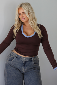 A woman wearing a dark brown, fitted, long sleeved, slightly cropped top with blue trim detailing at the v-neckline and at the sleeve hems, paired with dark acid washed barrel denim jeans, standing indoors in front of a white wall.