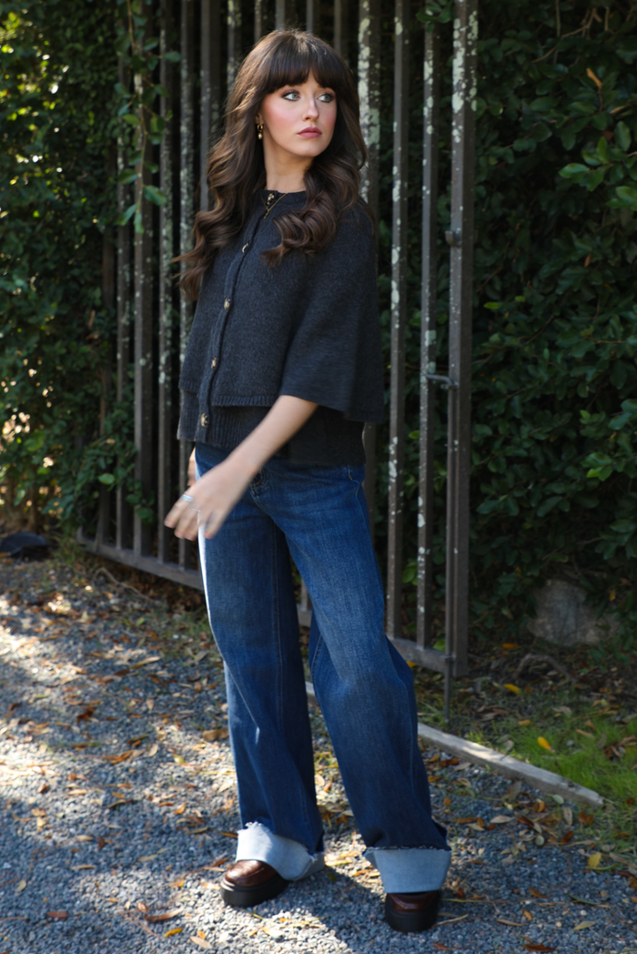 Woman model outdoors wearing a charcoal grey colored cape cardigan with functional button closures in the front and dark wash denim. 