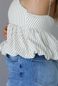 Woman wearing a white and taupe striped tank with medium wash denim shorts indoors against a white wall. The photo shows a closeup of the side of the top, which has a zipper closure. 