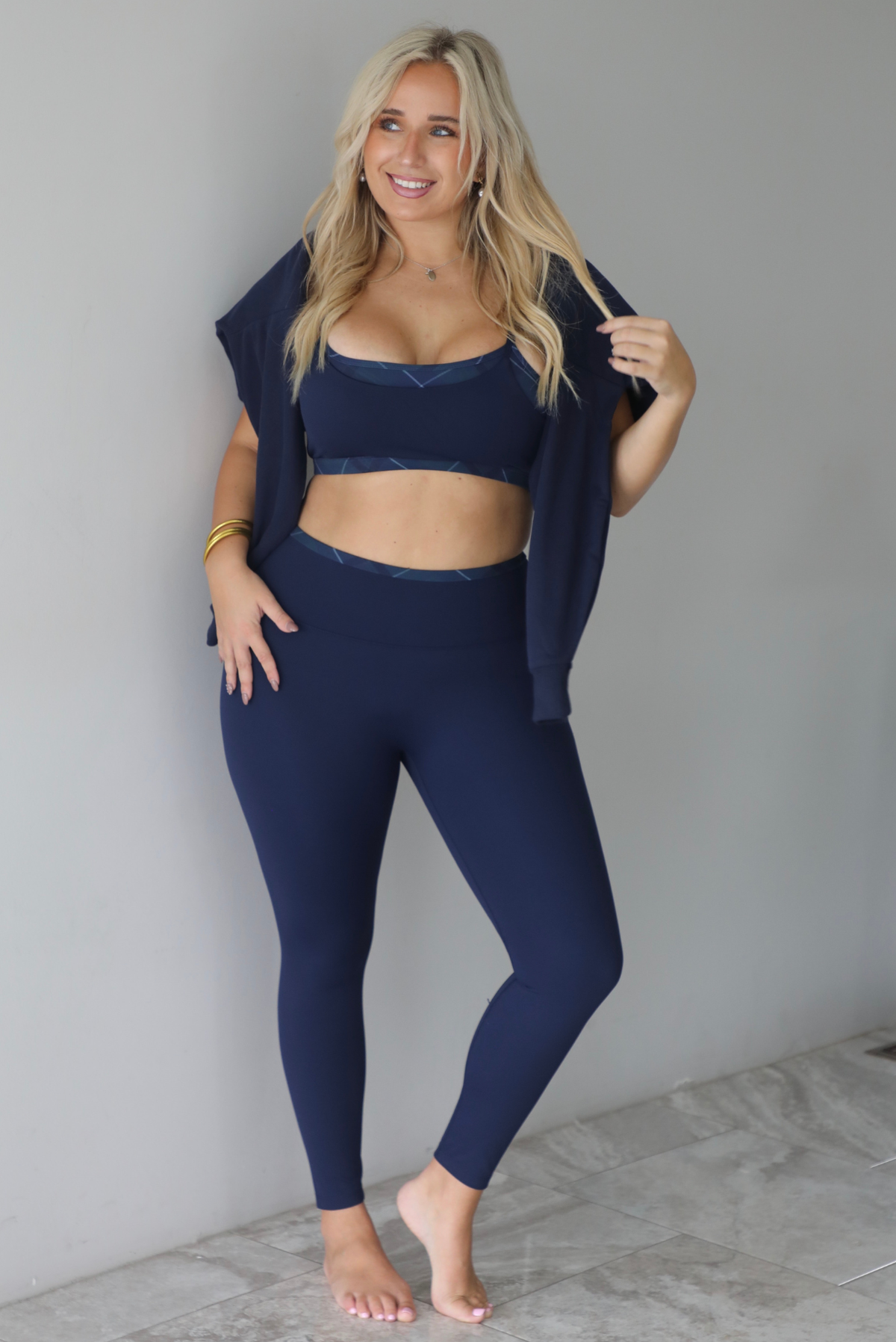 A woman wearing a navy sweater with Apreś written on the front in collegiate lettering in a plaid print over her shoulders in a draped detail, paired with a matching two piece navy set, standing indoors against a white wall with white marble flooring. 