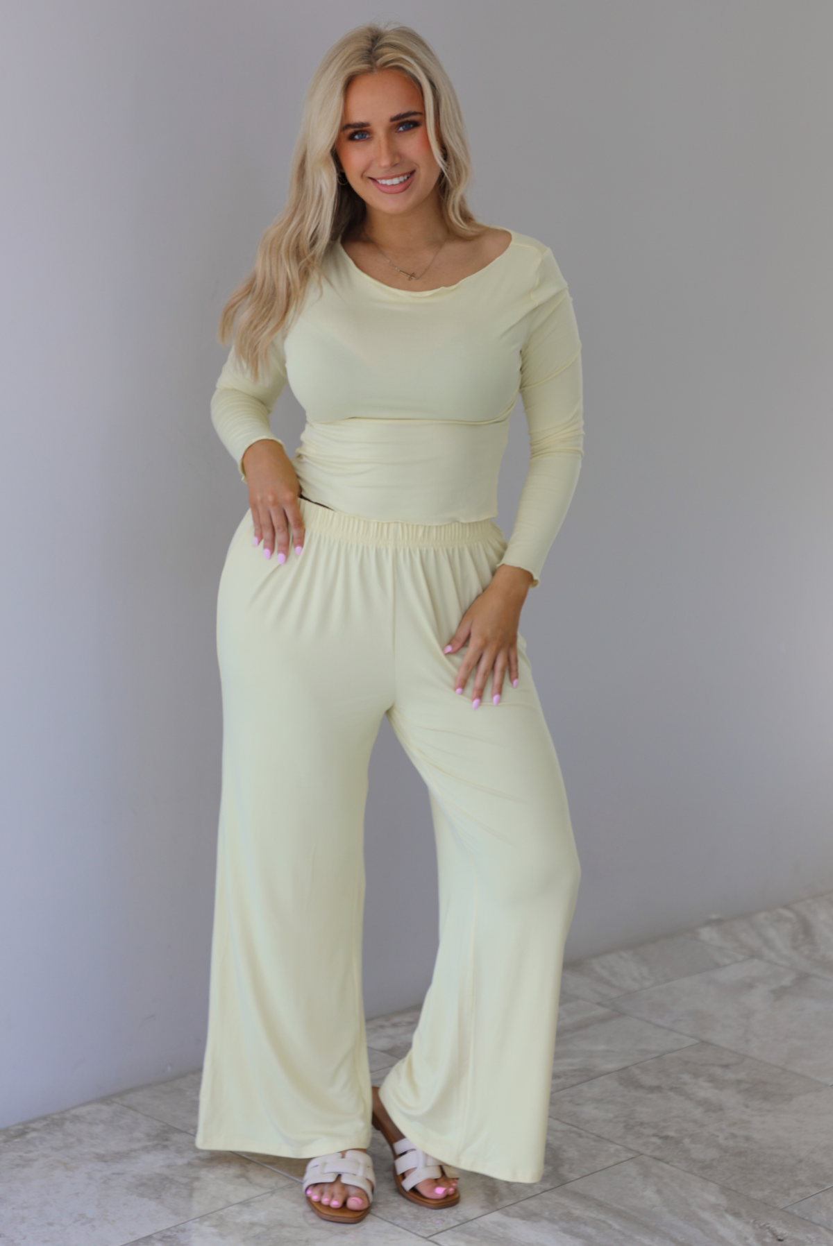 Woman wearing a light yellow three piece set featuring a long sleeved top, long, flared hem pants, and a long, matching cardigan. The photo is taken indoors against a white wall with white marble flooring. The photo shows the set without the cardigan. 