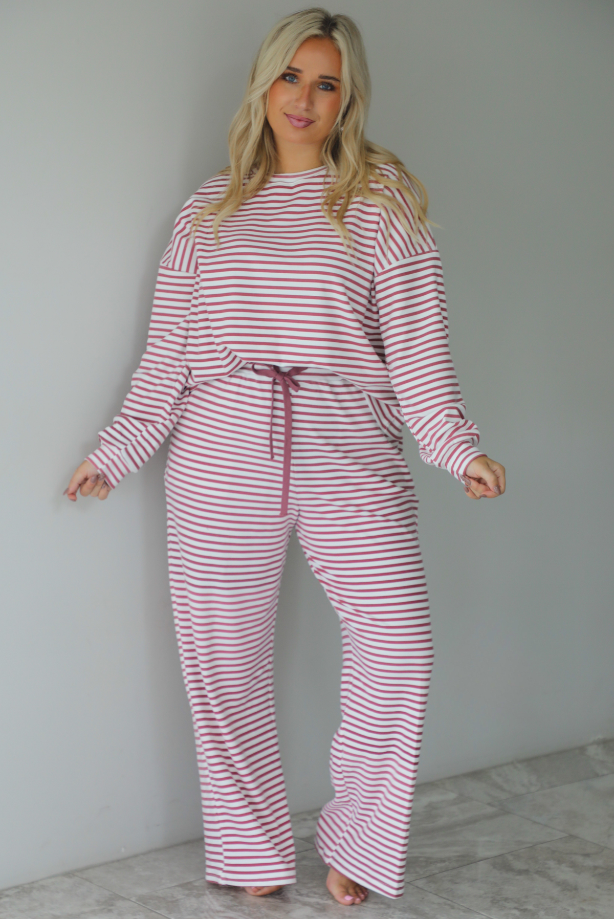 A woman wearing a matching two piece set featuring a long sleeved top and high waisted wide leg pants with a berry and white striped pattern throughout, standing indoors against a white wall with white marble flooring. 