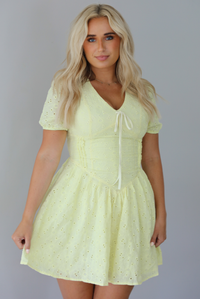 Woman wearing a light yellow mini dress with a floral eyelet design throughout, v-neckline, short sleeves, fitted drop waist bodice, and a flowy mini length skirt indoors against a white wall.