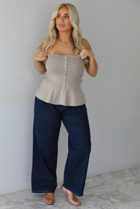A woman wearing a beige strapless button-front peplum top and dark denim wide-leg jeans, standing indoors against a white wall with white marble flooring. 