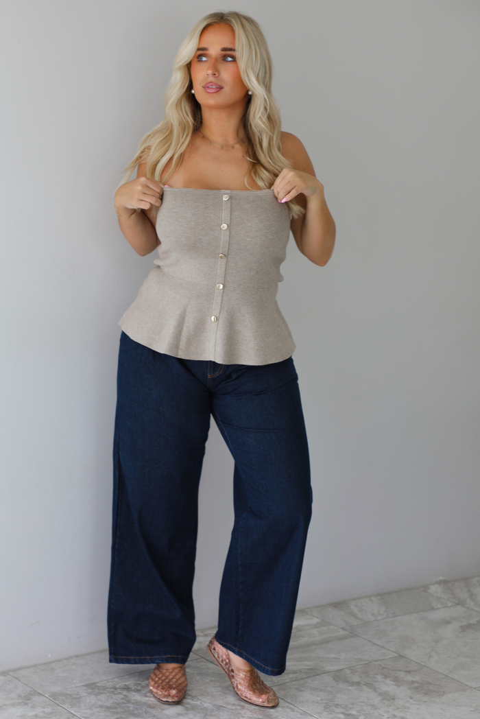 A woman wearing a beige strapless button-front peplum top and dark denim wide-leg jeans, standing indoors against a white wall with white marble flooring. 