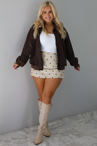 A woman wearing a brown suede jacket, paired with a white tank and cream colored mini skort with a brown polka dot print pattern throughout, and sand colored knee boots, standing indoors against a white wall with white marble flooring. 