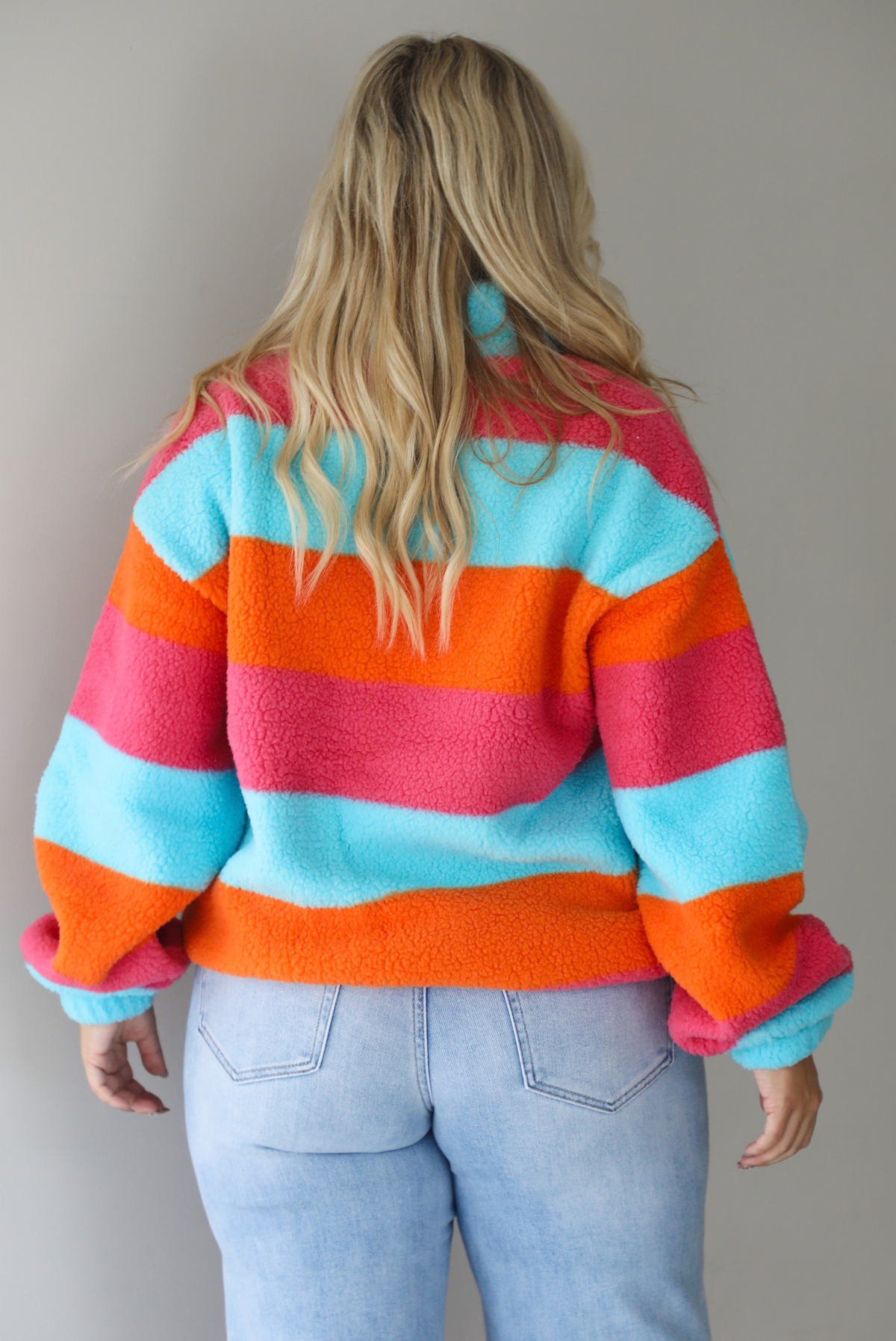 A woman shown from behind wearing a turquoise, pink, and orange striped sherpa quarter-zip sweater with light wash denim jeans, standing indoors against a white wall.