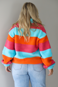 A woman shown from behind wearing a turquoise, pink, and orange striped sherpa quarter-zip sweater with light wash denim jeans, standing indoors against a white wall.