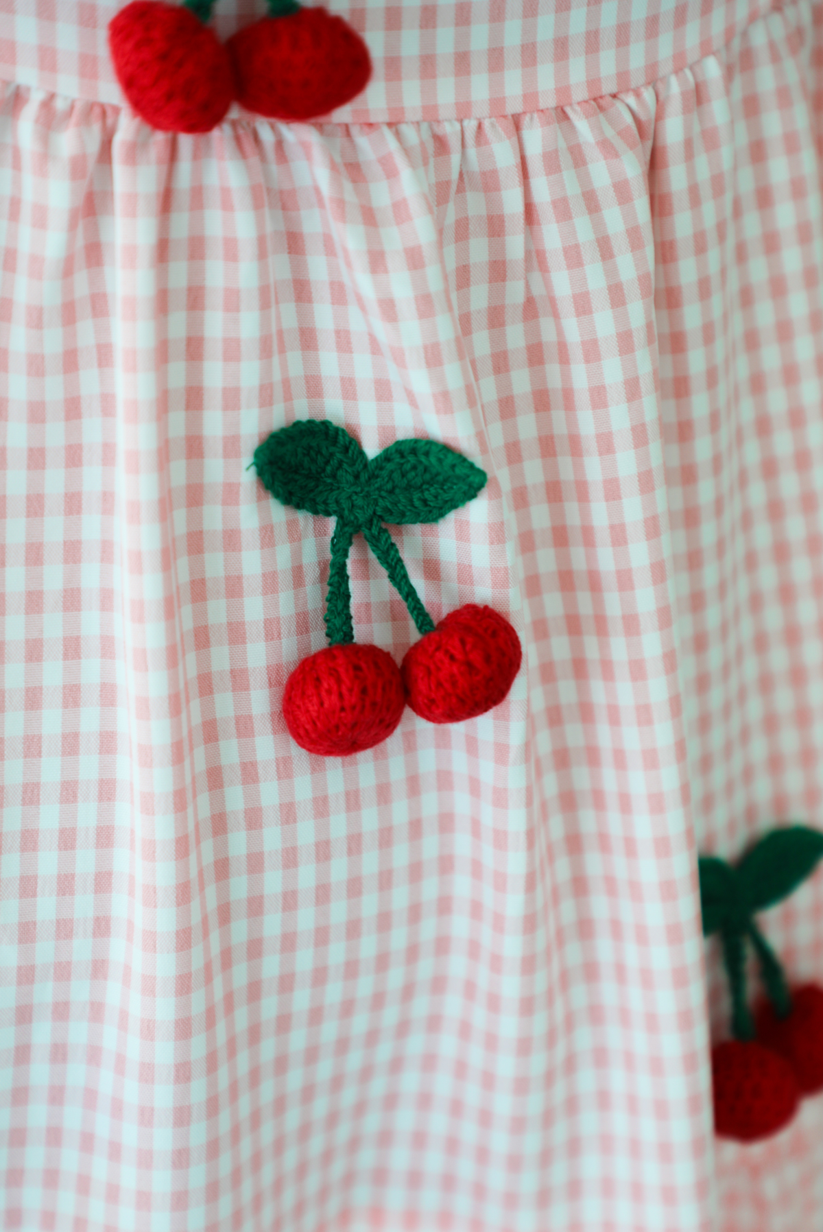 Woman wearing a pink and white gingham  maxi dress with thick straps, a fitted bodice, flowy skirt, and crocheted cherry details. The photo shows a closeup of the crocheted cherry detail, which connects to the dress with the leaves and dangles.