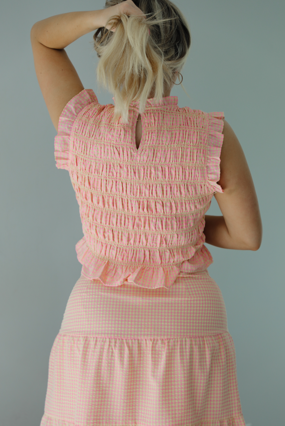Woman wearing a pink and pale yellow gingham print two piece set featuring a smocked top and high waisted, maxi length skirt. The photo is taken indoors against a white wall and shows a closeup of the back of the smocked top, which has a keyhole button closure at the top.