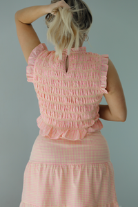 Woman wearing a pink and pale yellow gingham print two piece set featuring a smocked top and high waisted, maxi length skirt. The photo is taken indoors against a white wall and shows a closeup of the back of the smocked top, which has a keyhole button closure at the top.