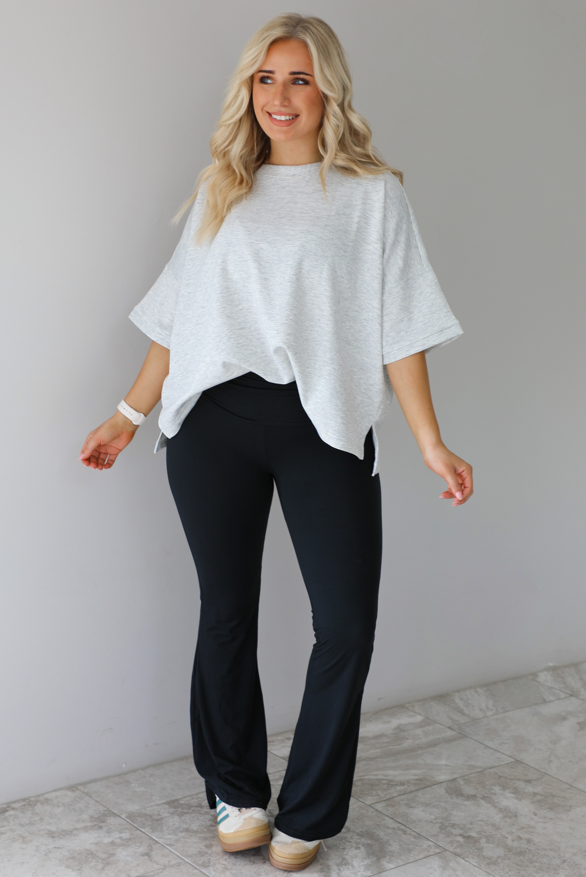 Woman wearing an oversized heather grey box tee with black leggings that flare slightly at the hems. The photo is taken indoors against a white wall with white marble flooring. 