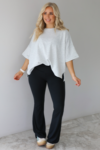 Woman wearing an oversized heather grey box tee with black leggings that flare slightly at the hems. The photo is taken indoors against a white wall with white marble flooring. 
