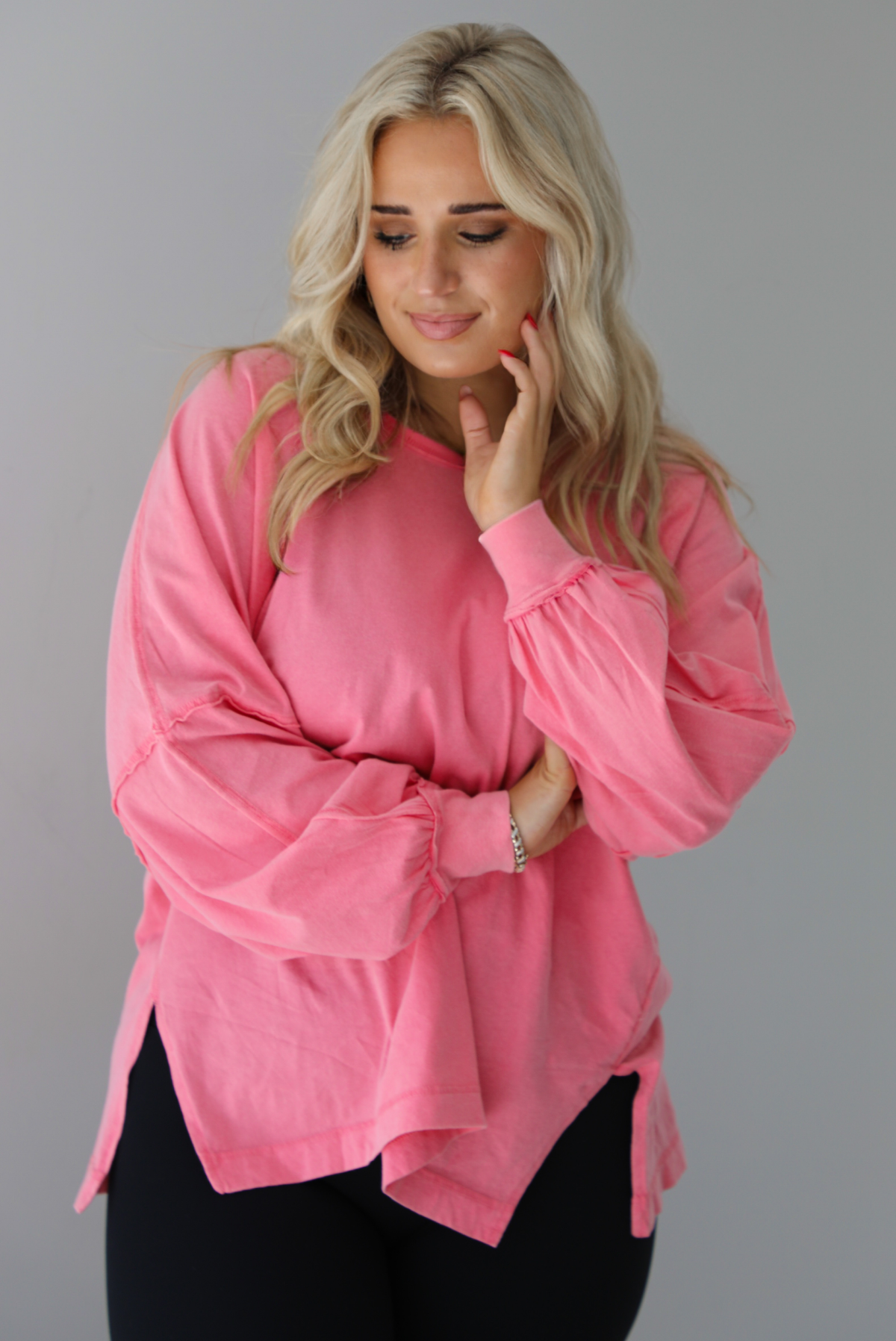 Woman wearing a pink oversized long sleeved top that has a high neckline, exposed seam details, split hem details on either side, and is lightweight. The top is modeled with a pair of fitted black leggings. The photo is modeled indoors against a white wall.