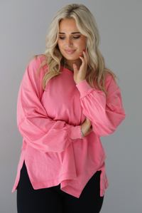 Woman wearing a pink oversized long sleeved top that has a high neckline, exposed seam details, split hem details on either side, and is lightweight. The top is modeled with a pair of fitted black leggings. The photo is modeled indoors against a white wall.