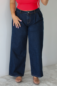 A woman wearing wide-leg dark denim jeans, a red top, and brown woven sandals, standing indoors against a white wall with white marble flooring. 