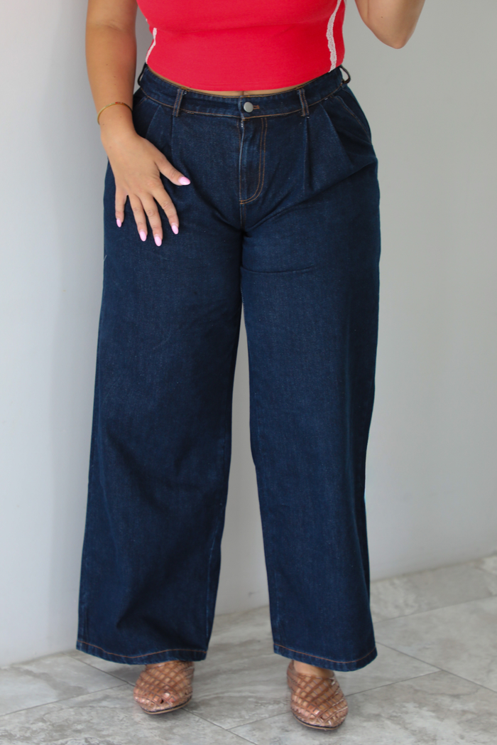 A woman wearing wide-leg dark denim jeans, a red top, and brown woven sandals, standing indoors against a white wall with white marble flooring. 