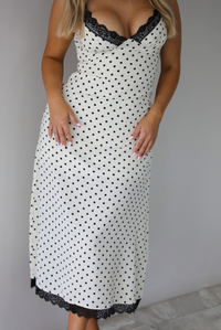 Woman wearing a white midi length dress with a black polka dot print pattern and black lace detailing at the v-neckline and bottom hem. The photo is taken indoors against a white wall with white marble flooring. 