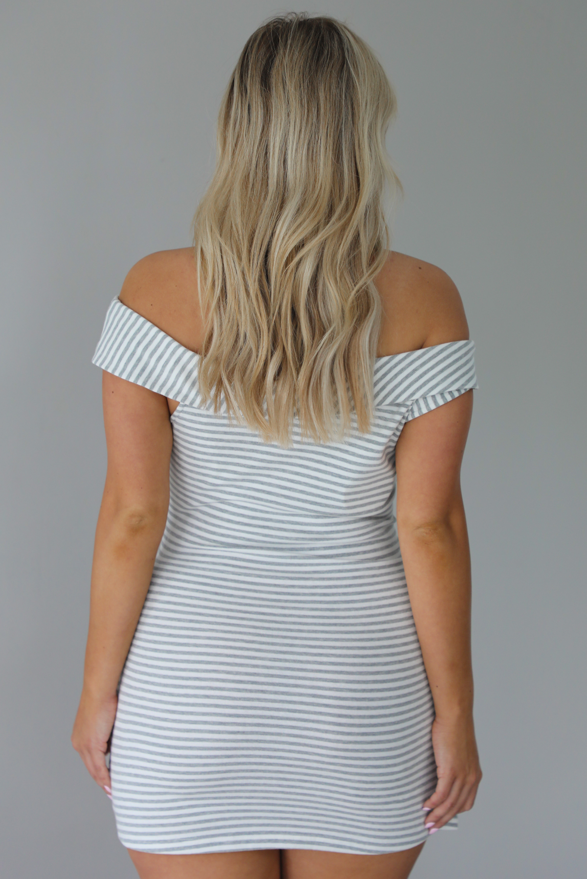Woman wearing an off the shoulder grey and white striped mini dress that has a tie detail in the front. The photo is taken indoors against a white wall and shows the back of the dress. 