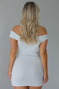 Woman wearing an off the shoulder grey and white striped mini dress that has a tie detail in the front. The photo is taken indoors against a white wall and shows the back of the dress. 