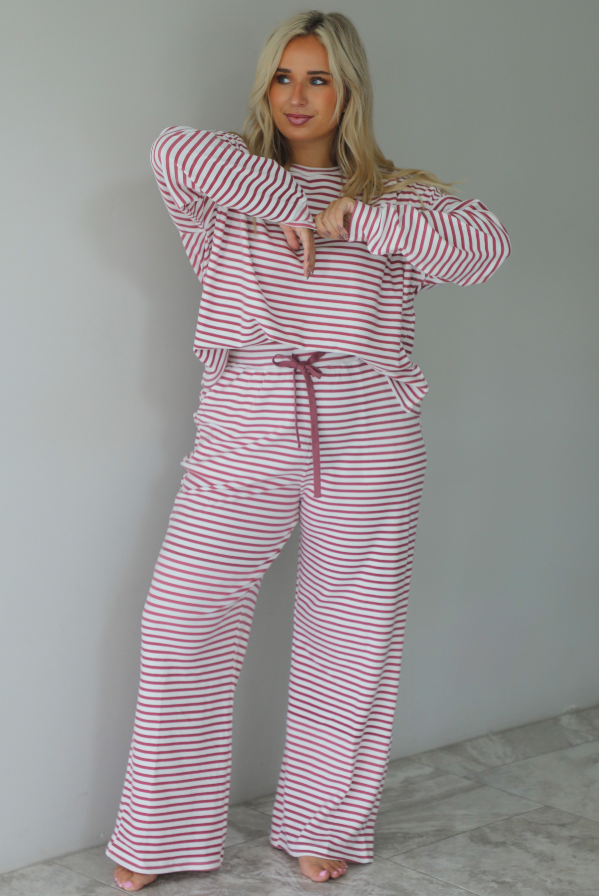 A woman wearing a matching two piece set featuring a long sleeved top and high waisted wide leg pants with a berry and white striped pattern throughout, standing indoors against a white wall with white marble flooring. 