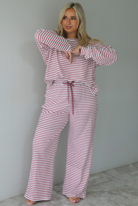 A woman wearing a matching two piece set featuring a long sleeved top and high waisted wide leg pants with a berry and white striped pattern throughout, standing indoors against a white wall with white marble flooring. 