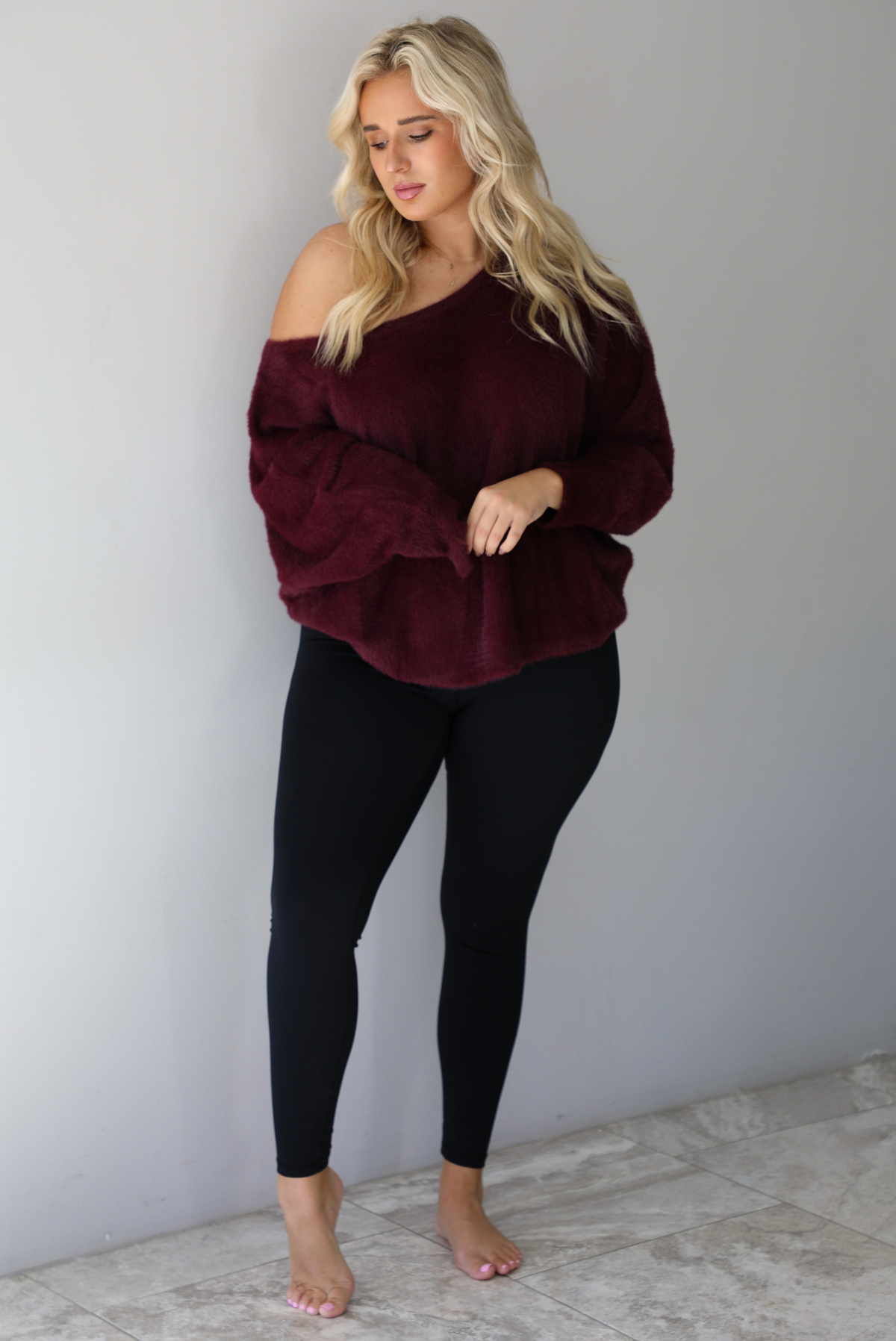 A woman wearing a maroon oversized long sleeved sweater, worn off the shoulder, paired with black leggings, indoors against a white wall with white marble flooring.
