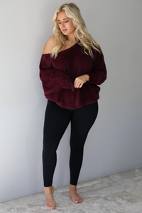 A woman wearing a maroon oversized long sleeved sweater, worn off the shoulder, paired with black leggings, indoors against a white wall with white marble flooring.