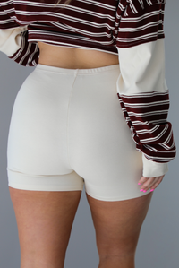 Woman wearing a burgundy and cream striped oversized collared top with cream colored biker shorts. The photo is taken indoors against a white wall. The photo shows a closeup of the back of the shorts. 