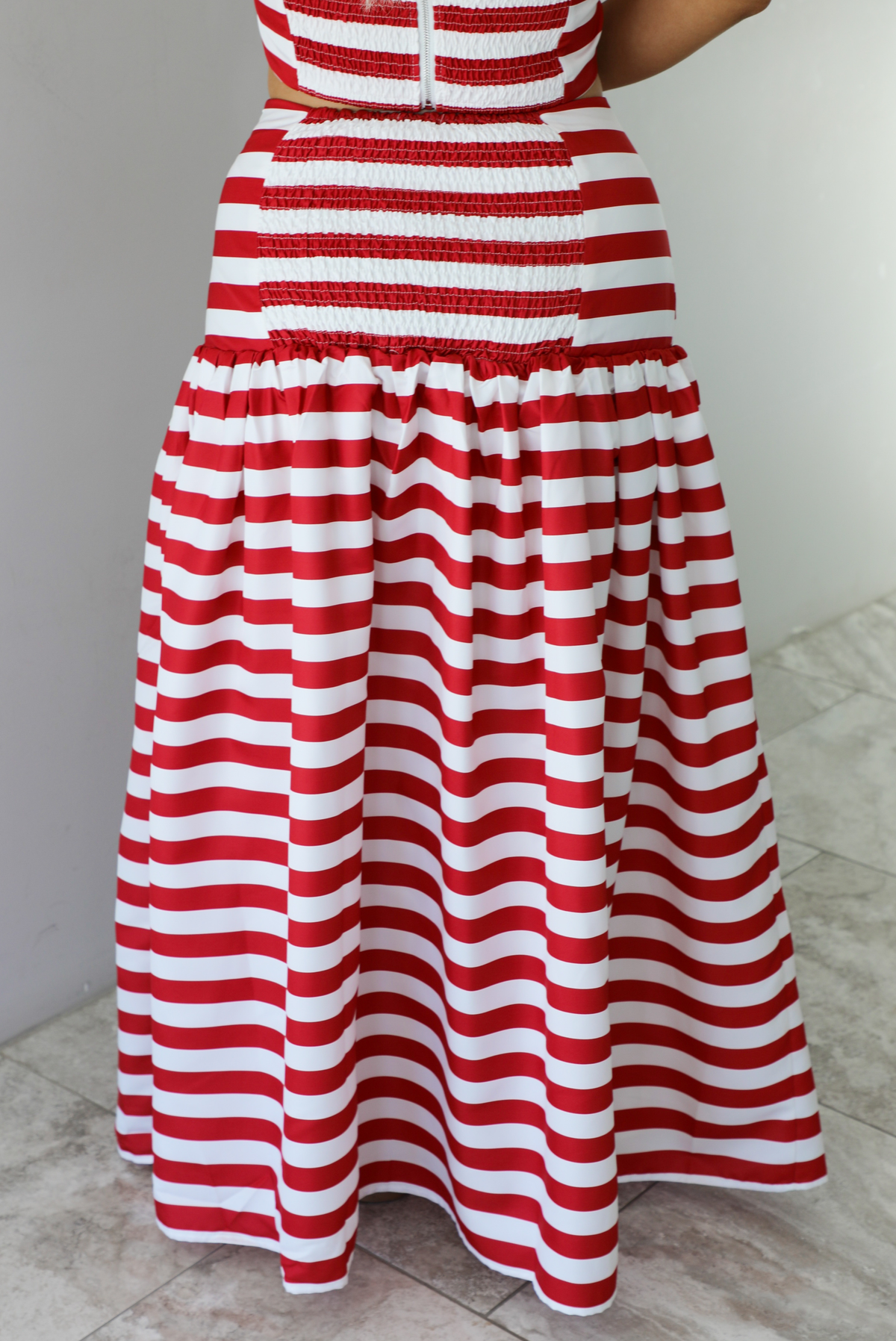 Woman wearing a red and white striped two piece set featuring a cropped top and maxi length, high waisted skirt. The photo is taken indoors against a white wall with white marble flooring, and shows a closeup of the back of the skirt, which has smocked detailing.