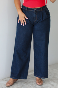 A woman wearing wide-leg dark denim jeans, a red top, and brown woven sandals, standing indoors against a white wall with white marble flooring. 