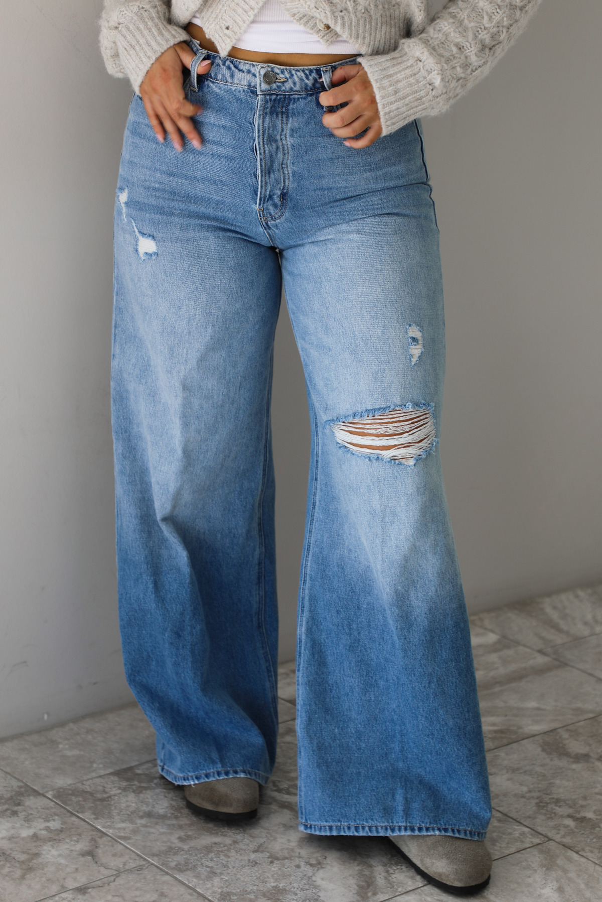 Woman wearing high waisted medium wash denim that have multiple button closures, functional belt loops, functional front and back pockets, and distressed details throughout with a flared hem. The photo is taken in front of a white wall and white marble flooring.