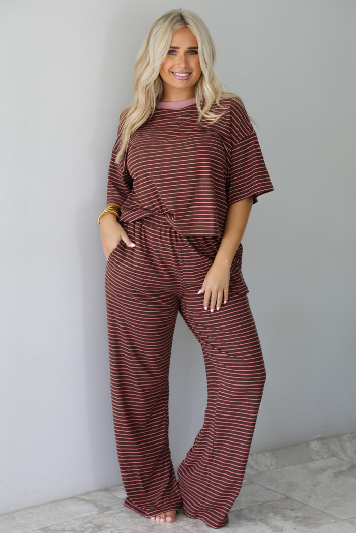 A woman wearing a brown and pink striped two piece set featuring a slightly cropped tee and high waisted pants, with the top slightly tucked into the pants, standing indoors against a white wall with white marble flooring. 