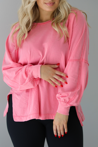 Woman wearing a pink oversized long sleeved top that has a high neckline, exposed seam details, split hem details on either side, and is lightweight. The top is modeled with a pair of fitted black leggings. The photo is modeled indoors against a white wall.