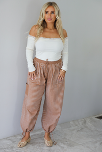 A woman wearing a cream off the shoulder long sleeved top tucked into a pair of camel and cream striped pants, standing indoors against a white wall with white marble flooring. 