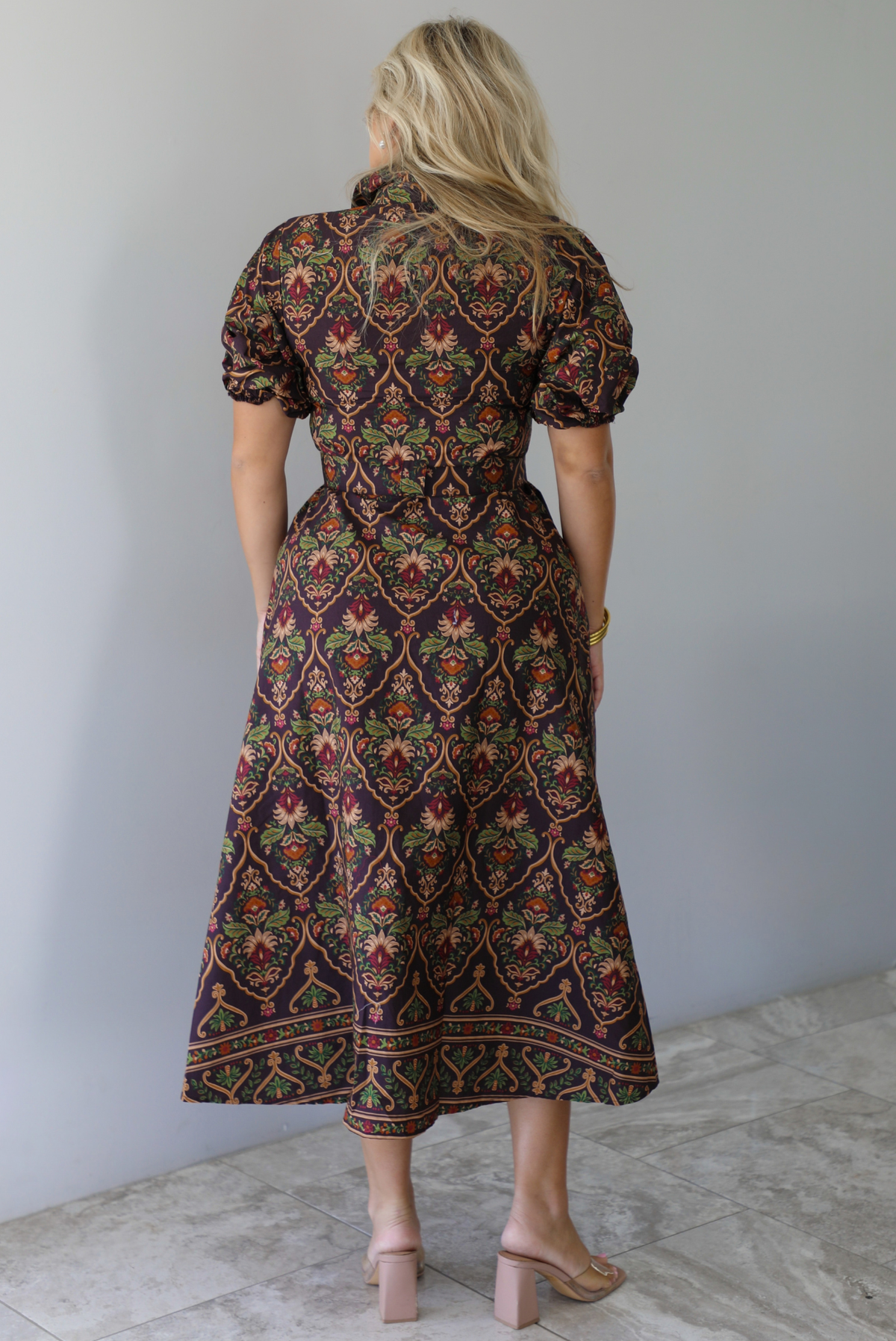 A woman shown from behind wearing a dark brown midi dress with a multi-colored design throughout, a collared neckline, short puffed sleeves, functional button closures along the front, a belted detail at the waistline, standing indoors against a white wall with white marble flooring. 