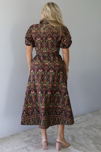 A woman shown from behind wearing a dark brown midi dress with a multi-colored design throughout, a collared neckline, short puffed sleeves, functional button closures along the front, a belted detail at the waistline, standing indoors against a white wall with white marble flooring. 