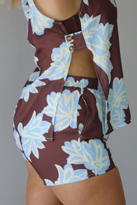 Woman wearing a brown, blue, and off white floral two piece set featuring a vest style top with functional button closures in the front and shorts. The photo is taken in front of a white wall and shows the side of the set, which has an open design and button closures on the side, and also shows the elastic waistline and functional pocket on the shorts. 