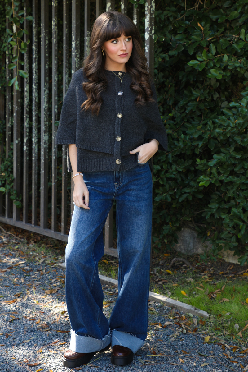 Woman model outdoors wearing a charcoal grey colored cape cardigan with functional button closures in the front and dark wash denim. 